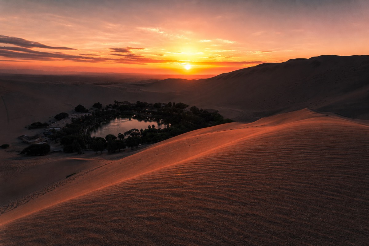 The Huacachina desert oasis lit up at dusk with its lagoon reflecting the surrounding dune-top buildings